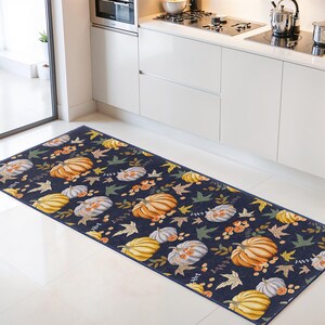 May include: A rectangular kitchen rug with a navy blue background. The rug features a repeating pattern of pumpkins in shades of orange, yellow, and white, along with green and brown leaves. The rug is placed on a white tile floor.