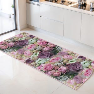 Könnte beinhalten: Ein rechteckiger Küchenteppich mit floralem Design. Der Teppich zeigt ein Muster aus rosa und lila Rosen mit grünem Laub auf einem hellrosa Hintergrund. Er liegt vor einer weißen Küchenzeile.