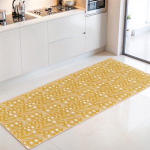 May include: A rectangular kitchen rug with a repeating pattern of white leaves and berries on a mustard yellow background. The rug is placed on a white tile floor in front of a white kitchen cabinet.