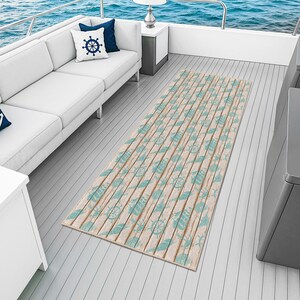 Puede incluir: Una alfombra rectangular de temática náutica con un patrón de vetas de madera clara. La alfombra presenta un diseño repetido de conchas marinas azul claro, timones y otros elementos náuticos. La alfombra está colocada en la cubierta de un barco con un sofá blanco y cojines azules.