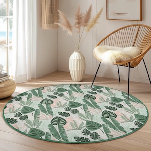 May include: A round rug with a tropical leaf pattern in shades of green, pink, and white. The rug has a green border and is placed on a wooden floor. The rug is approximately 60 inches in diameter.