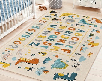 May include: A rectangular beige rug with a dinosaur and alphabet design. The rug features colorful illustrations of dinosaurs, the alphabet, and numbers. The border has a yellow outline. The rug is placed in a nursery setting.