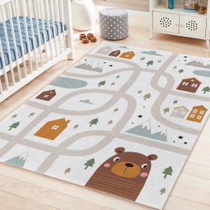 May include: A rectangular children's rug with a road map design. The rug features a light gray background with roads, houses, trees, mountains, and a brown bear. The houses are in shades of brown and orange, and the trees are green.