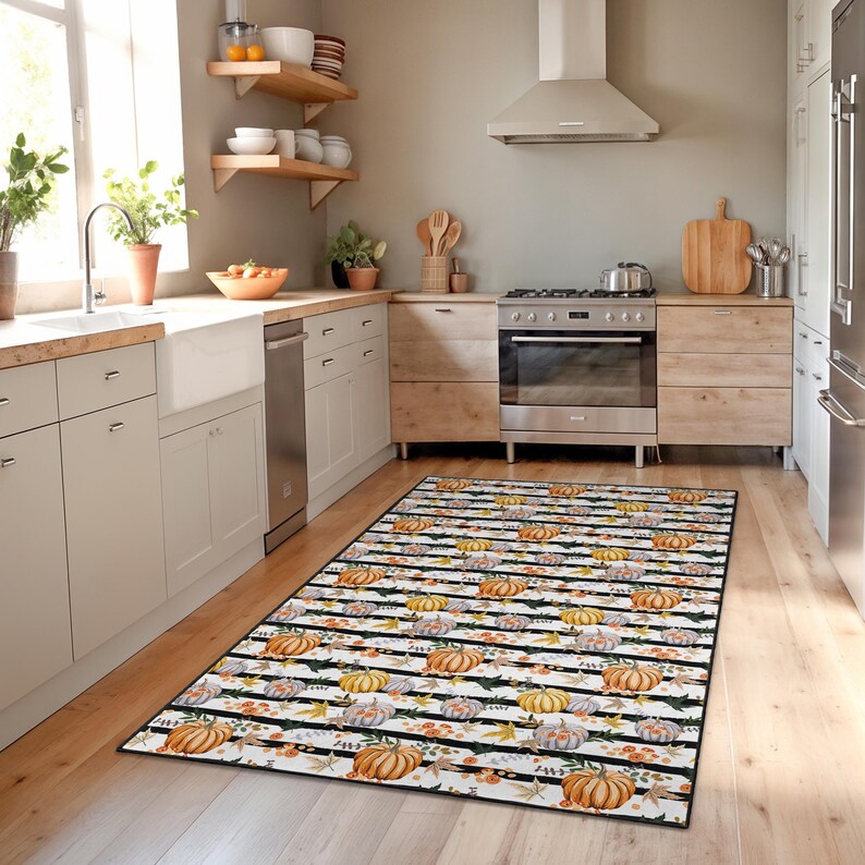 May include: A rectangular kitchen rug with a black and white striped pattern. The rug features a repeating design of pumpkins in shades of orange, yellow, and grey, along with autumn leaves. The rug is placed on a wooden floor in a kitchen setting.