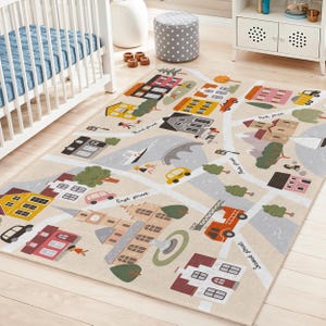 May include: A rectangular beige rug with a colorful town map design. The rug features roads, buildings, trees, and vehicles. Streets are labeled "First Street", "Second Street", and "Third Street". The rug is placed on a light wood floor.