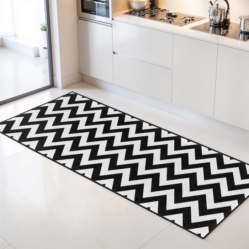 May include: A black and white chevron patterned kitchen rug. The rug is rectangular and lies on a white tile floor. The zig-zag pattern is a classic design element, adding a modern touch to the kitchen space. The rug is placed in front of the kitchen cabinets.