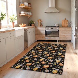 Puede incluir: Una alfombra rectangular negra con un patr&oacute;n repetido de calabazas, hojas y bayas en tonos naranja, amarillo y blanco. La alfombra est&aacute; colocada sobre un suelo de madera en una cocina.