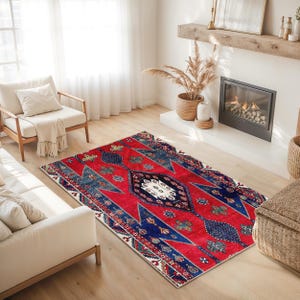 May include: A rectangular red rug with a central diamond pattern and geometric designs in blue, white, and green. The rug is placed on a light-colored wooden floor in a living room setting.