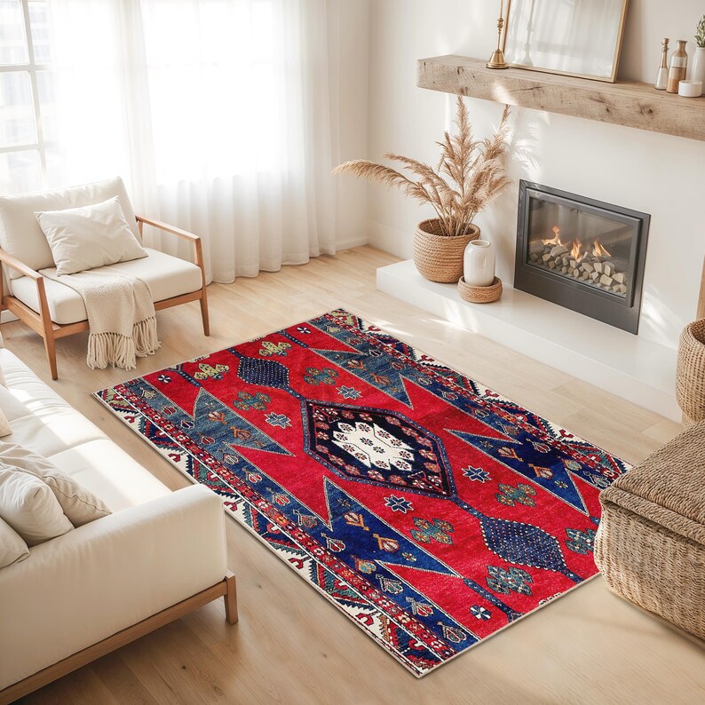 May include: A rectangular red rug with a central diamond pattern and geometric designs in blue, white, and green. The rug is placed on a light-colored wooden floor in a living room setting.