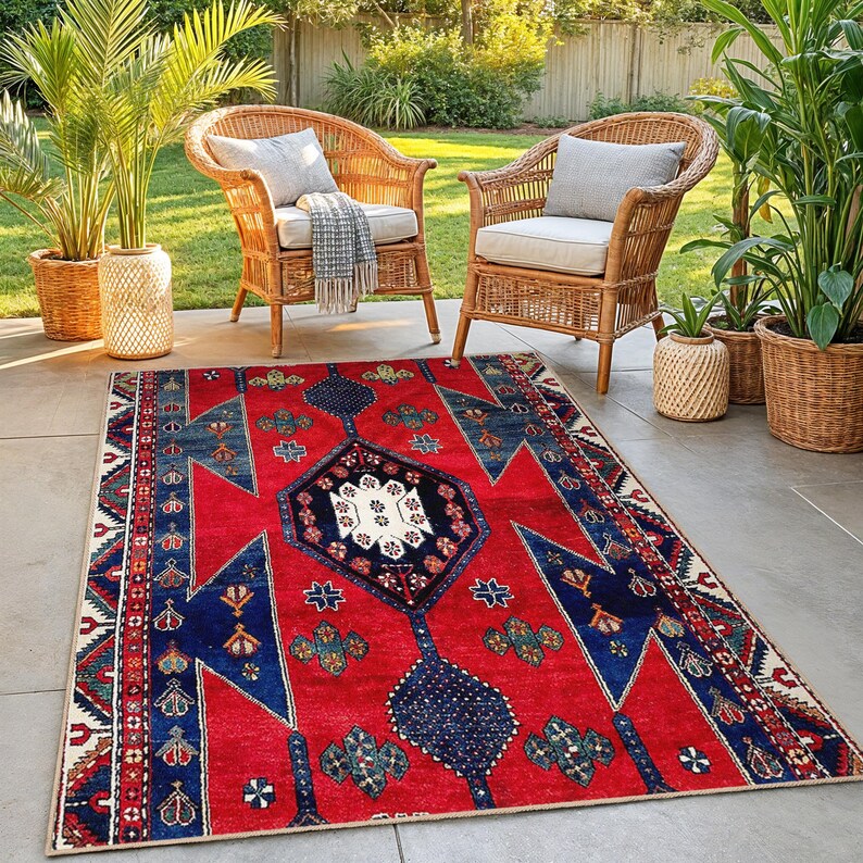 May include: A vibrant red area rug with a geometric design, featuring navy blue and white accents. The rug is placed on a patio with wicker chairs and potted plants, creating an inviting outdoor living space.
