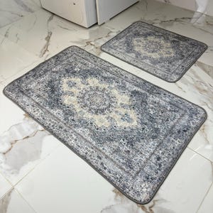 Op de afbeelding: Twee gepatroneerde badmatten in grijstinten en wit. De grotere mat heeft een ingewikkelder ontwerp met een centraal medaillon. De kleinere mat heeft een eenvoudiger ontwerp.