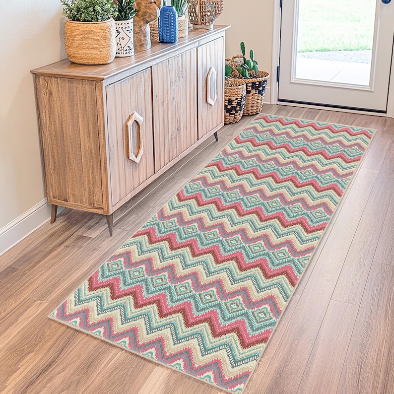 May include: A rectangular area rug with a colorful zigzag pattern in shades of pink, teal, cream, and brown. The rug is placed on a wooden floor in front of a wooden cabinet. The rug's design features a repeating geometric pattern.