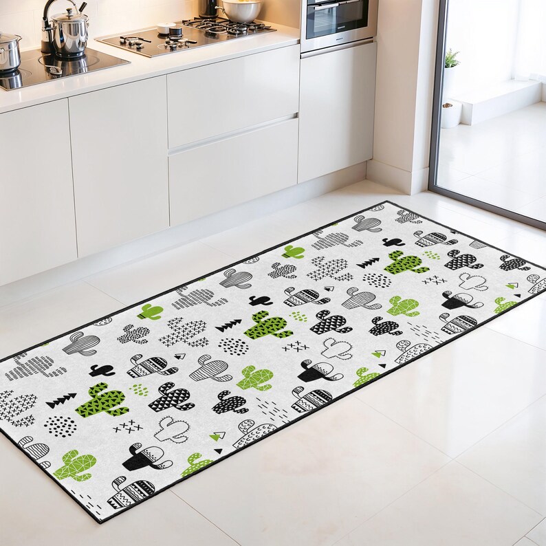 May include: A rectangular kitchen rug with a white background and a black border. The rug features a repeating pattern of stylized cacti in black, gray, and green. The rug is placed in front of a white kitchen cabinet.
