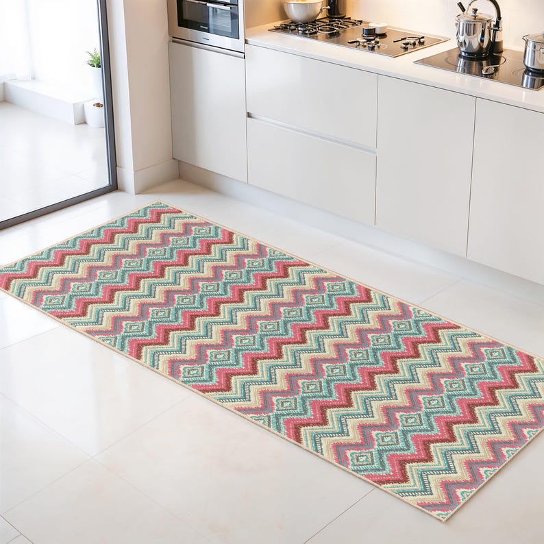 May include: A rectangular kitchen runner rug with a repeating zigzag pattern in shades of pink, teal, beige, and brown. The rug is placed on a white tile floor in front of white kitchen cabinets.