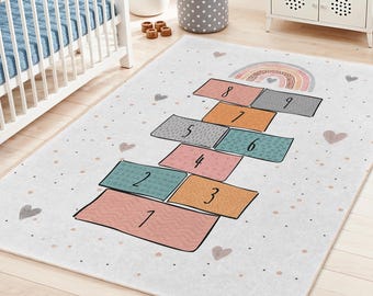 May include: A rectangular children's rug with a hopscotch design. The rug features colorful squares numbered 1 through 9, a rainbow, and heart shapes on a white background. The rug is designed for a nursery or playroom.