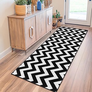 May include: A black and white chevron pattern runner rug. The rug is rectangular and placed on a hardwood floor. The rug is in front of a wooden cabinet and a white door.