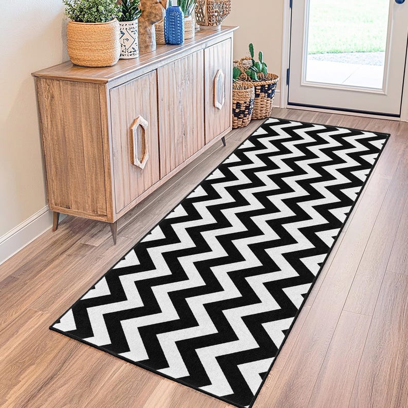May include: A black and white chevron pattern runner rug. The rug is rectangular and placed on a hardwood floor. The rug is in front of a wooden cabinet and a white door.