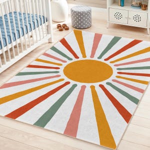May include: A rectangular rug with a sunburst design. The rug features a large yellow circle in the centre, with colourful rays of orange, red, pink, green, and yellow radiating outwards against a white background. The rug is placed on a light-coloured wooden floor.
