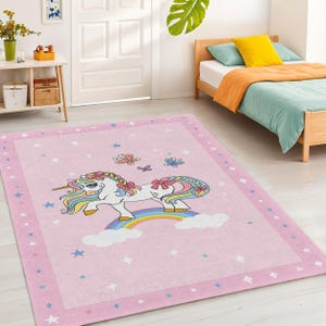 Può includere: Tappeto rettangolare rosa con un design a unicorno dei cartoni animati. L'unicorno ha una criniera e una coda arcobaleno, e si trova su un arcobaleno. Il tappeto ha un bordo rosa con stelle e farfalle, ideale per la cameretta dei bambini. Misura circa 150 cm x 200 cm.