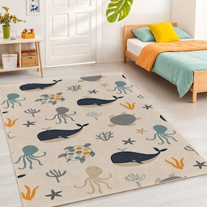 Puede incluir: Alfombra rectangular beige con un patrón repetido de vida marina. El diseño incluye ballenas azul marino, pulpos turquesa, tortugas grises, algas naranjas y estrellas de mar. La alfombra está en una habitación con una cama y una mesita auxiliar.