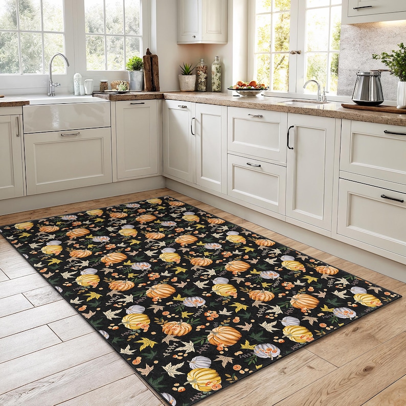 Puede incluir: Una alfombra rectangular negra con un patr&oacute;n repetido de calabazas, hojas y bayas en tonos naranja, amarillo y verde. La alfombra est&aacute; colocada en una cocina con gabinetes blancos y un suelo de madera.