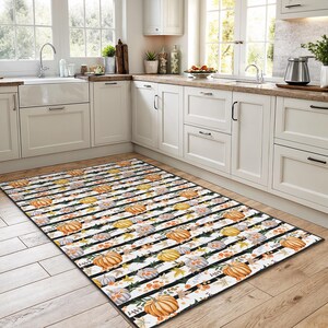 May include: A rectangular kitchen rug with a repeating pattern of pumpkins in orange, yellow, and grey, with floral and leaf accents. The design is set against a white background with black stripes.