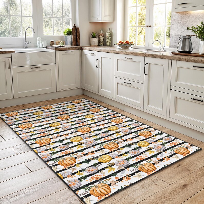 May include: A rectangular kitchen rug with a repeating pattern of pumpkins in orange, yellow, and grey, with floral and leaf accents. The design is set against a white background with black stripes.