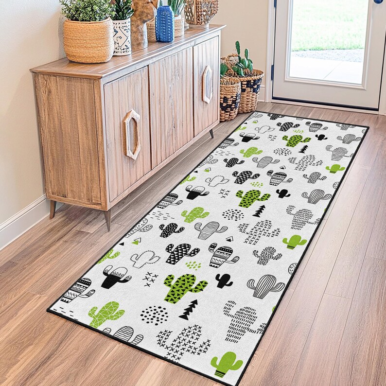 May include: A rectangular white rug with a black border featuring a repeating pattern of black and green cacti in various designs. The rug is placed on a wooden floor near a wooden cabinet and a white door.