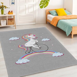 Può includere: Un tappeto rettangolare grigio con un design stravagante di un unicorno bianco che va in bicicletta su un arcobaleno. L'unicorno ha una criniera e una coda colorate. Il tappeto include anche nuvole e stelle.