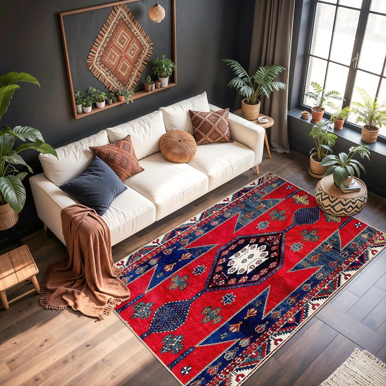 May include: A vibrant red and blue area rug with a geometric pattern. The rug features a central diamond design with intricate details, surrounded by bold red and blue shapes. The rug is placed in a living room setting with a white sofa and plants.
