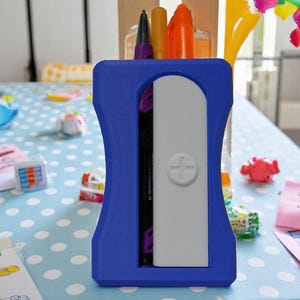 Könnte beinhalten: Ein blauer Plastik-Bleistifthalter mit einem weißen Fach und einem Pluszeichen. Der Halter ist mit Stiften und Markern gefüllt.