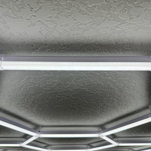 May include: An overhead view of a white and gray tent frame. The frame is constructed of white, rectangular tubes connected by clear plastic joints. The tent's interior is a textured gray surface, and the frame is illuminated by white lights.