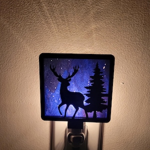 May include: A nightlight with a blue and white starry sky background. The nightlight features a silhouette of a deer and a pine tree.