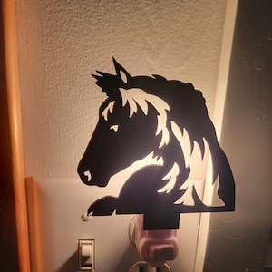May include: A black horse silhouette nightlight plugged into a white electrical outlet with a white light switch. The nightlight is lit and casts a warm glow.
