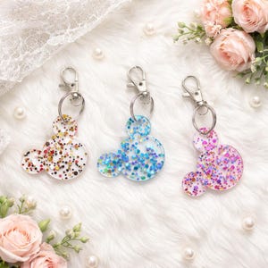 May include: Three clear acrylic keychains in a mouse shape, each with a different color of glitter. One has red, gold, and black glitter, another has blue glitter, and the third has pink and purple glitter. Each keychain has a silver clasp.
