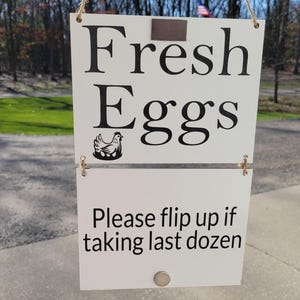 May include: A white sign with the words "Fresh Eggs" in large black letters, with a hen and eggs graphic. Below, it reads "Please flip up if taking last dozen." The sign hangs by twine.
