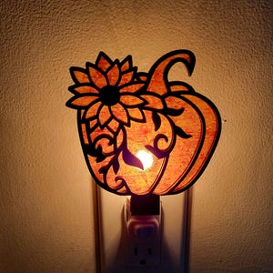 May include: A decorative night light shaped like a pumpkin with a sunflower design. The pumpkin is orange with a black outline, and the sunflower is black. The night light is plugged into a wall outlet and emits a warm glow.