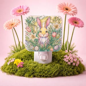 May include: A decorative night light featuring a watercolor illustration of a bunny surrounded by flowers and Easter eggs. The night light is set in a bed of green moss, with pink and yellow flowers in the background.