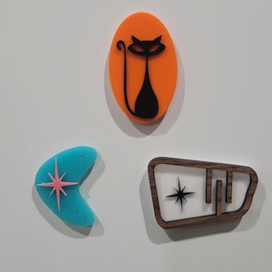 May include: Three retro-style magnets. A turquoise boomerang with a pink starburst, an orange oval with a black cat silhouette, and a brown wooden shape with a star and geometric design.