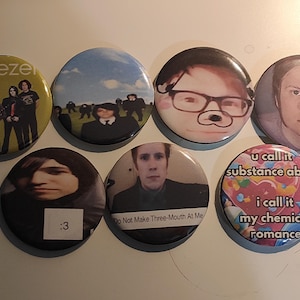 May include: A collection of seven round pinback buttons. One button features the band Weezer. Other buttons have images of people, and text that reads "Do Not Make Three-Mouth At Me", and "u call it substance abus i call it my chemical romance".