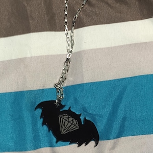 Peut inclure: Un collier à chaîne argentée avec un pendentif noir en forme de chauve-souris orné d'un diamant. Le fond est un tissu rayé marron, blanc et bleu.