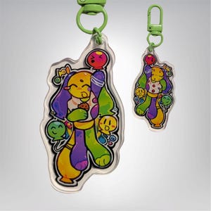Puede incluir: Dos llaveros de acrílico transparente con diseños de personajes de dibujos animados. El llavero más grande presenta un personaje amarillo con cabello morado, sosteniendo una galleta. El llavero más pequeño tiene un diseño similar. Ambos tienen herrajes verdes.