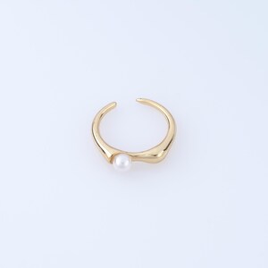 May include: A gold-toned open ring featuring a smooth, curved band with an irregular, organic shape. A single, small, round white pearl is set at one end of the opening, creating a contrast between the metallic gold finish and the pearl.