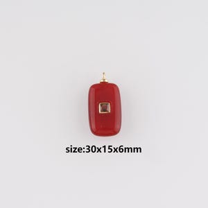 Puede incluir: Un colgante rojo con una anilla dorada y un acento cuadrado de color dorado. El colgante es rectangular con bordes redondeados. El texto "size:30x15x6mm" es visible. El colgante mide aproximadamente 3,0 x 1,5 x 0,6 cm.