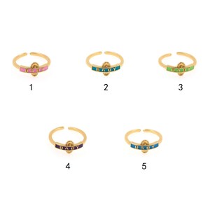 May include: Five gold rings with the word "BABY" in white lettering on a colored enamel band. The rings are numbered 1 through 5 and each ring has a different color band: pink, teal, green, purple, and blue.