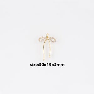 May include: A gold-coloured bow-shaped pendant with a loop at the top. The bow is decorated with small, clear gemstones. The pendant measures 30x19x3mm.