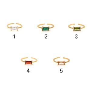 May include: Five gold rings with different colored gemstones. The rings are all the same style, with a thin band and a rectangular gemstone in the center. The gemstones are clear, green, dark green, red, and light brown.
