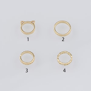 Könnte beinhalten: Vier goldfarbene Ringe werden vor einem hellgrauen Hintergrund gezeigt. Ein Ring hat Katzenohren mit rosa Akzenten, ein anderer hat ein schlichtes Band, und zwei haben strukturierte Designs. Die Ringe sind mit 1 bis 4 nummeriert.