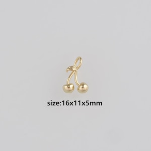 Puede incluir: Un charm de cereza dorado con dos esferas redondas y un tallo curvo. El charm mide aproximadamente 16x11x5mm. El texto "size: 16x11x5mm" es visible debajo del charm.