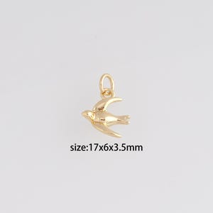 Può includere: Un ciondolo a forma di uccello dorato con un anello per appenderlo. Il ciondolo è in posizione di volo, con le ali spiegate. Il testo "size: 17x6x3.5mm" è sotto.